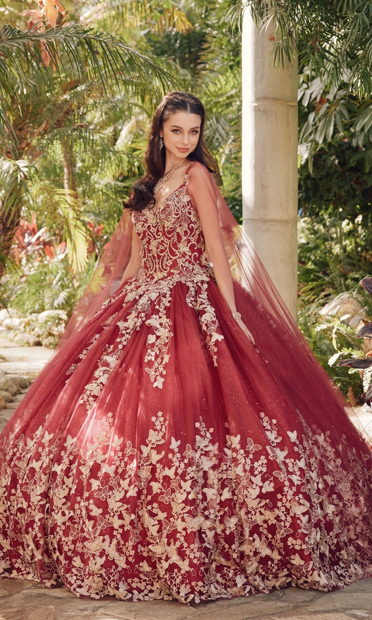 A woman stands outdoors in the Juliet 1442 Quinceañera Dress with Sheer Cape, a voluminous red ball gown featuring intricate white floral embroidery, surrounded by lush greenery.